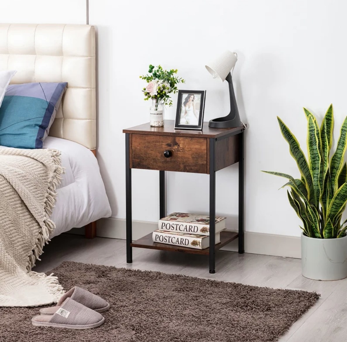 Wooden bedside table with lamp, picture frame, flowers on top, midde drawer, and books on bottom shelf