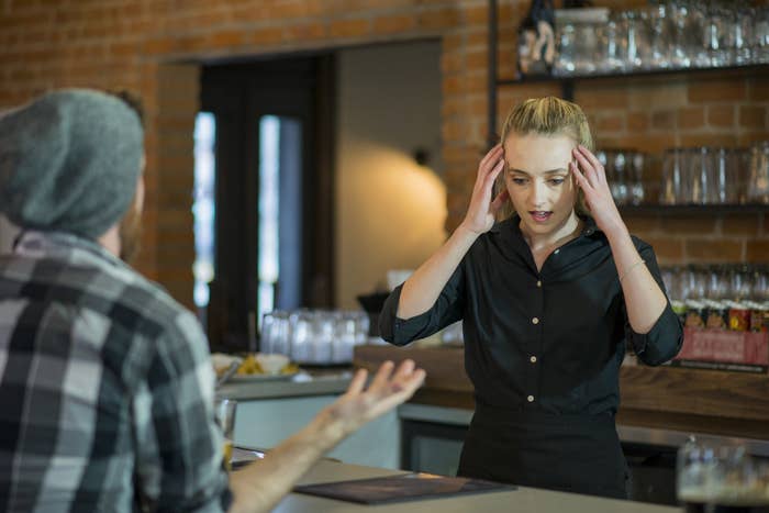 Stressed female young adult waitress holding her head in stress as someone complains to her behind the bar of a restaurant. She is wearing a black button up.