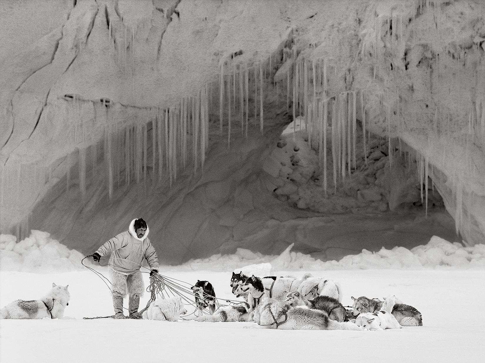 A man in traditional arctic dress with a lot of sled dogs sitting in front of a cave