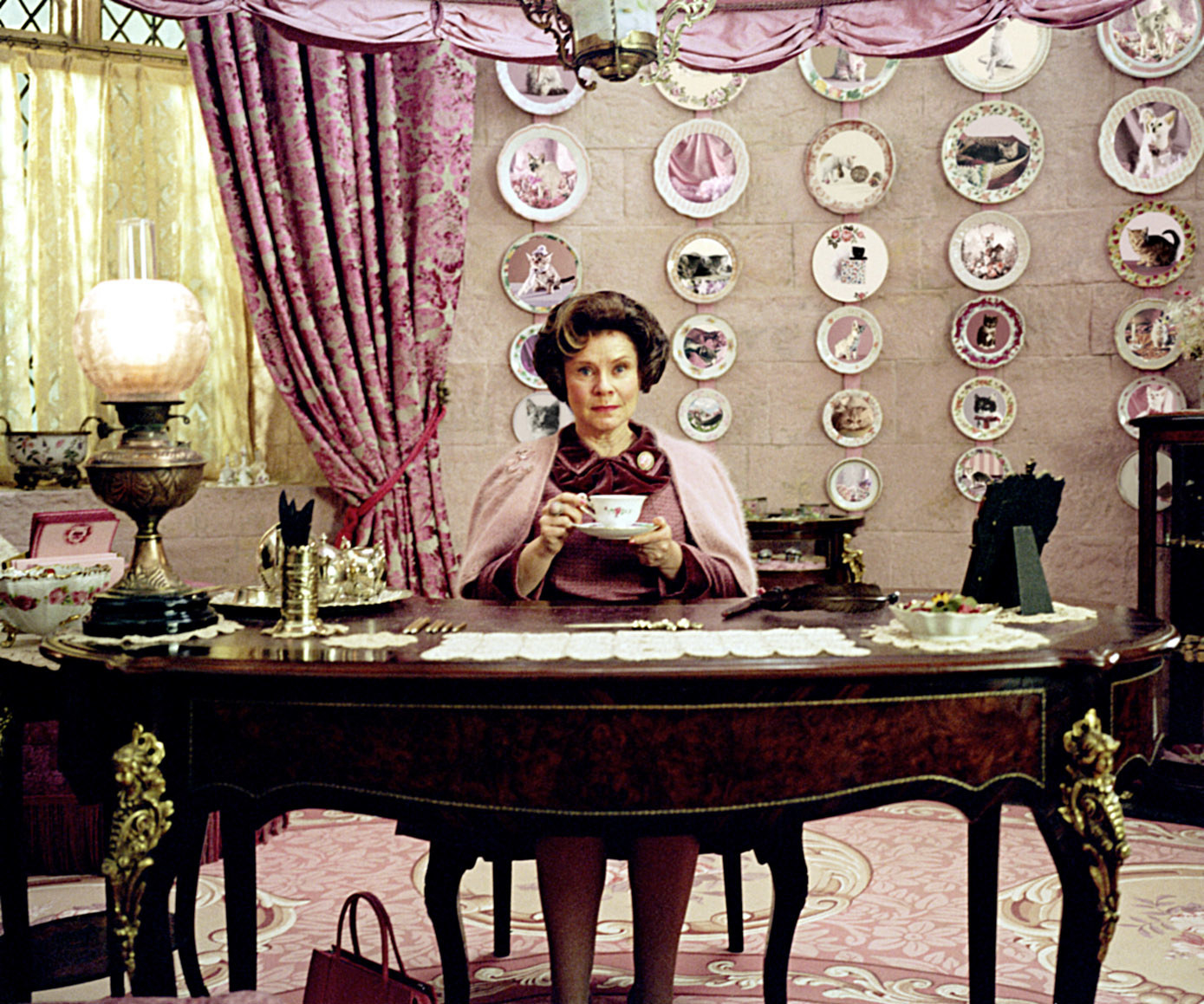 Dolores drinking tea in her office that&#x27;s entirely pink, has antique furniture, and tons of decorative plates with cats on them