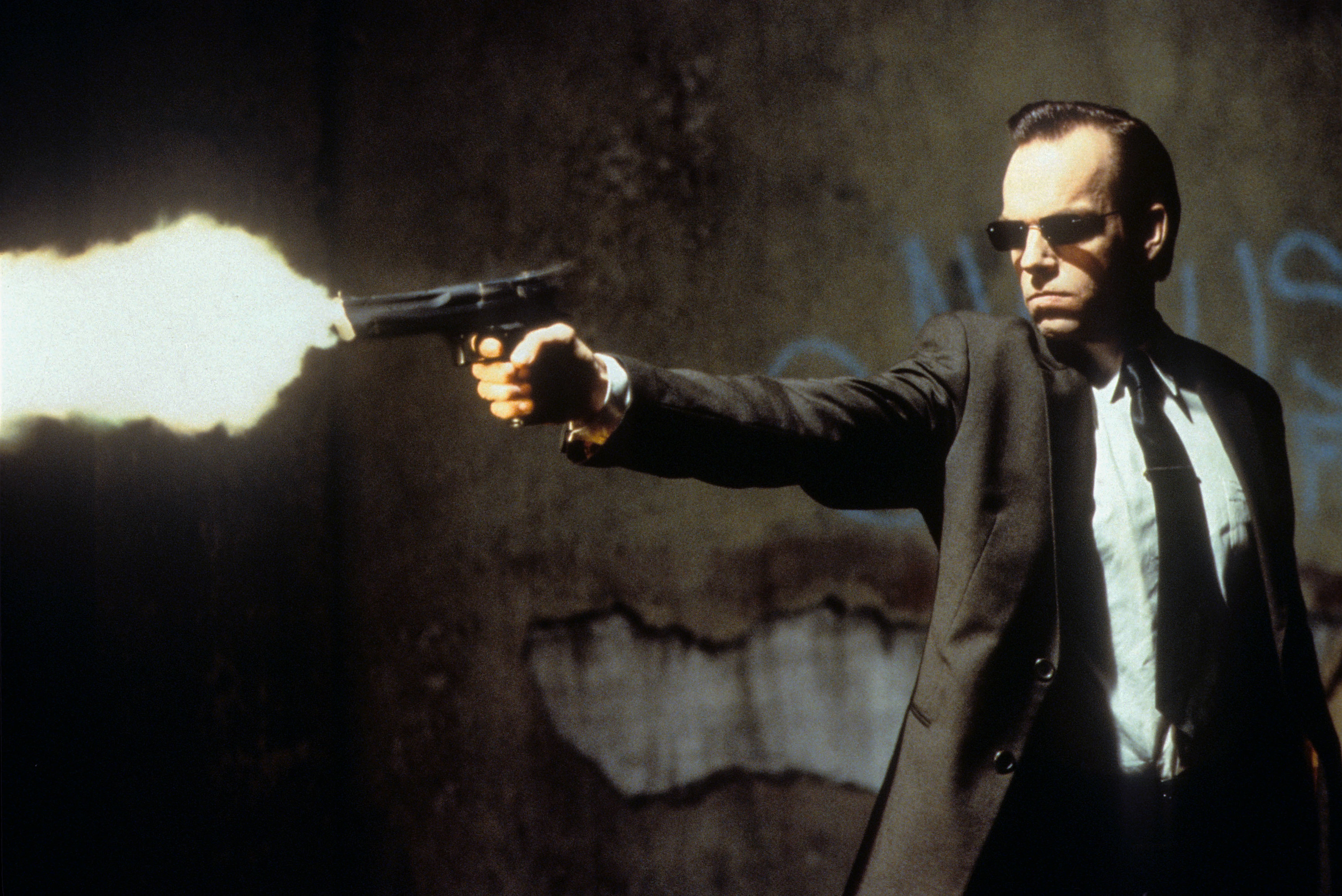 Agent Smith shooting a gun, wearing his usual suit and sunglasses