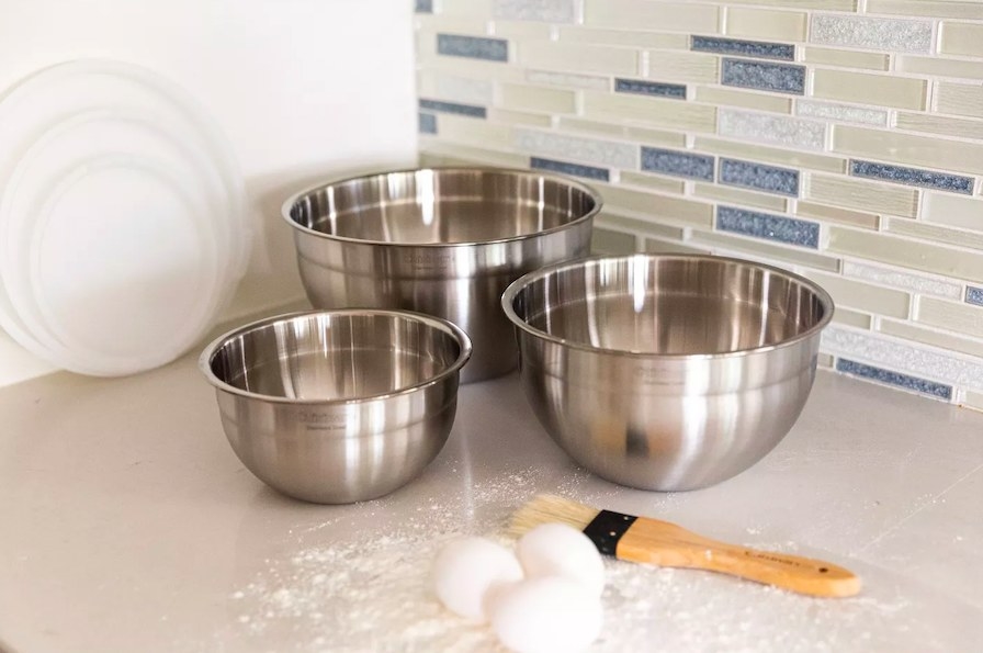 Three stainless steel bowls shown on a countertop with eggs, flour a pastry brush and their lids