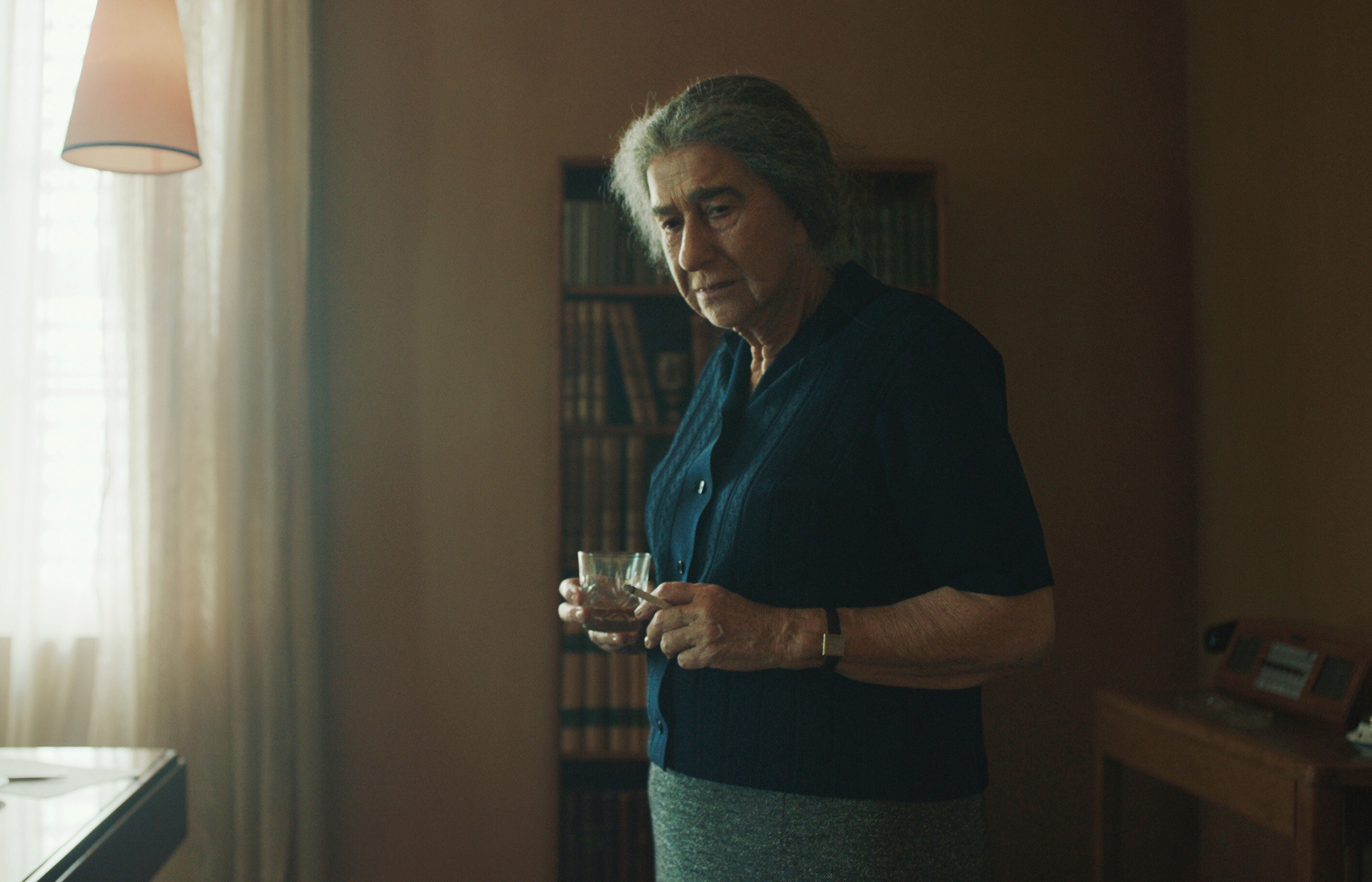 Helen as Golda standing in a room and holding a glass and a cigarette