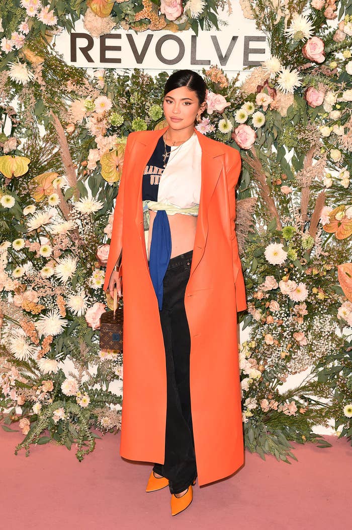 Kylie poses for a photo in front of a wall of flowers and the word REVOLVE