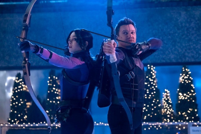 Clint Barton and Kate Bishop standing alongside each other in matching purple outfits, aiming their bows and arrows off-screen