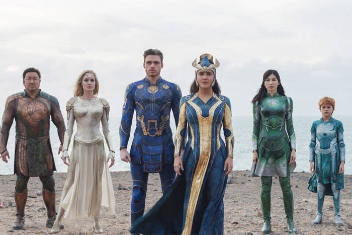 The characters Gilgamesh, Thena, Ikaris, Ajak, Sersi, and Sprite standing in a line on a beach in "Eternals", wearing their team outfits
