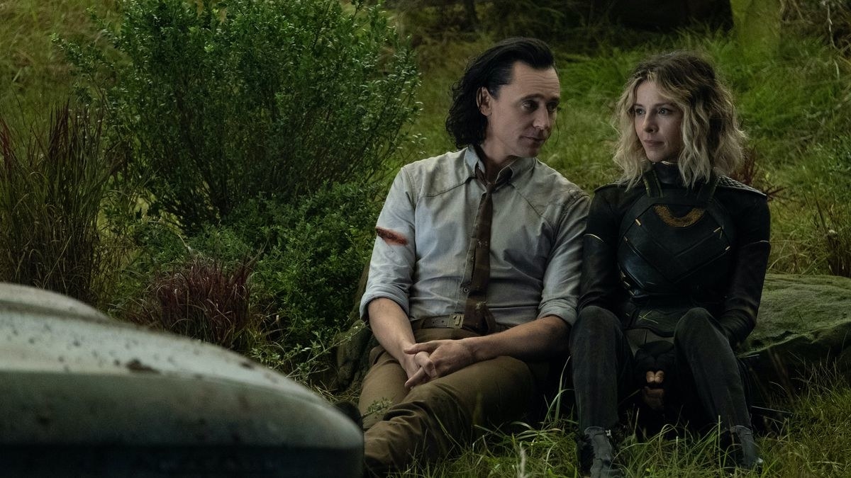 Loki in a dirty, torn shirt with a bloodstain on the right arm, sat on the grass next to Sylvie in a green and gold leather outfit by some plants