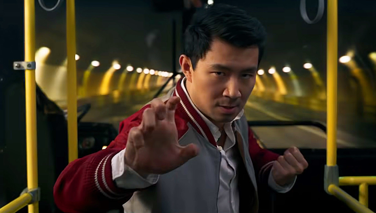 Shang-Chi standing on a bus in a grey and red jacket, holding his hands up in a fighting position