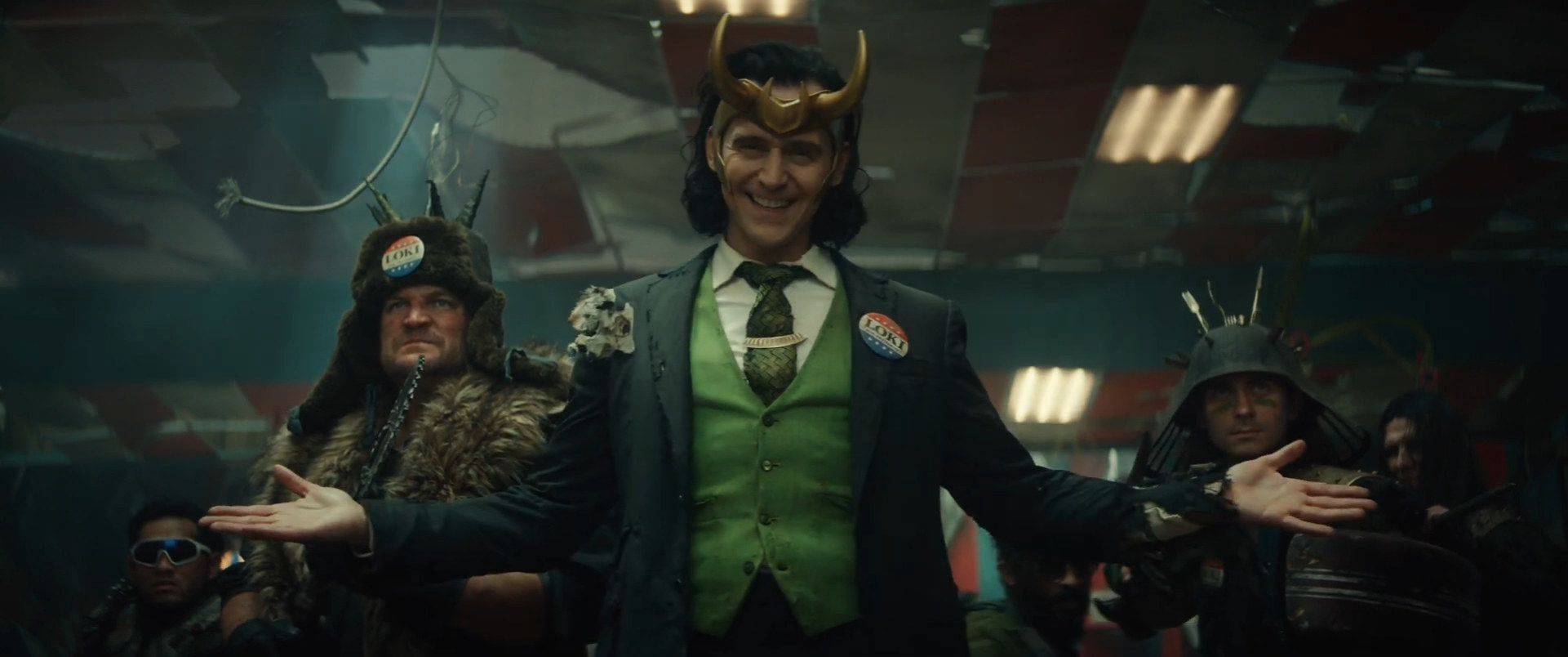 President Loki in a green suit and tie, wearing gold horns and smiling with his arms outstretched, while some of his goons stand behind him wearing horned hats
