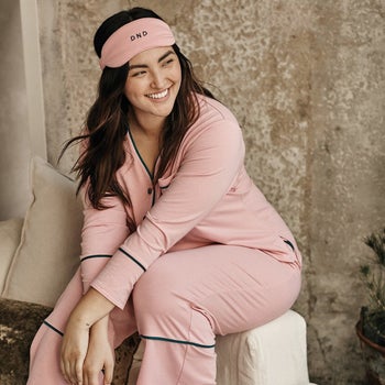 model sitting on a cushion wearing pink pajamas with black piping and matching eye mask
