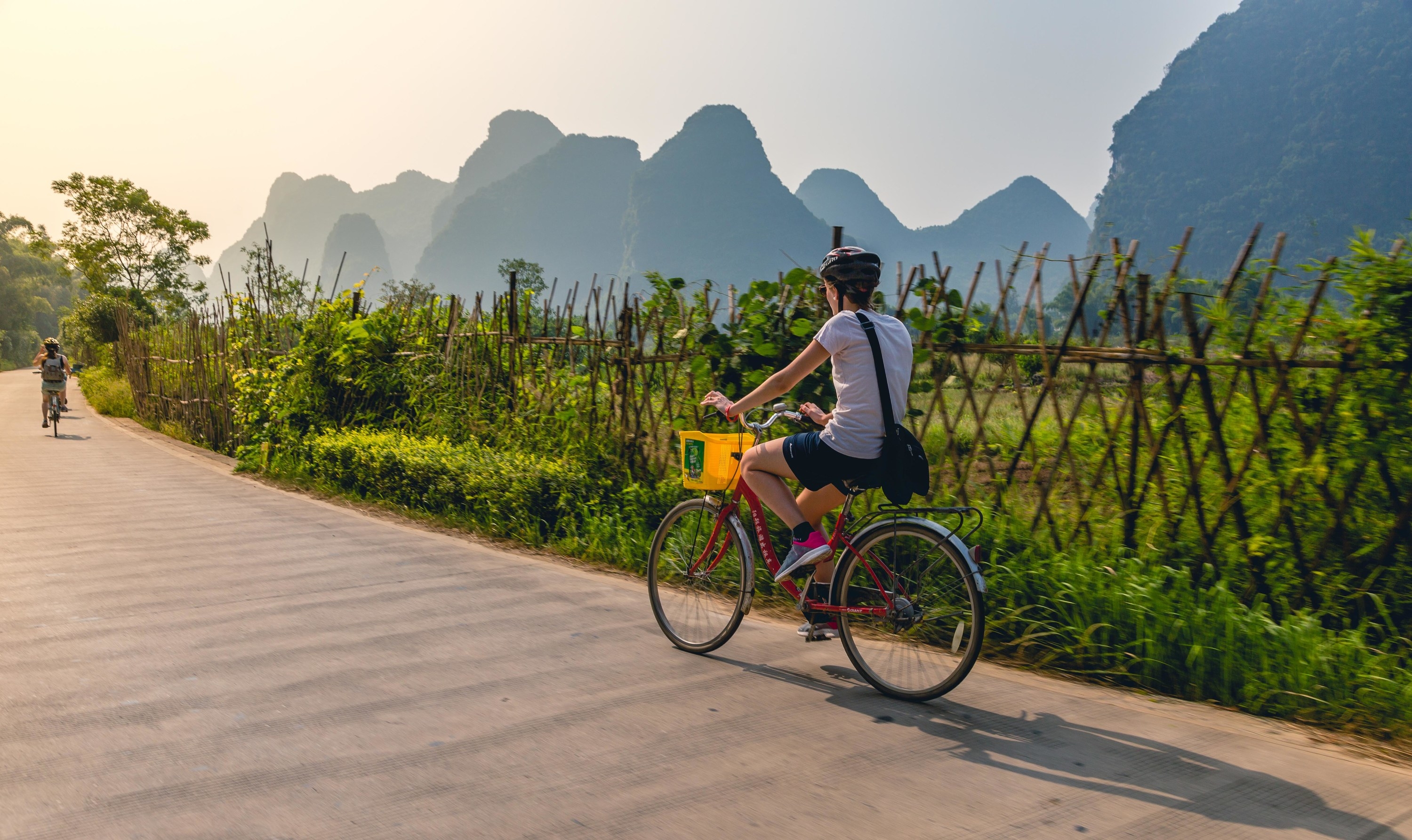 a woman riding a bicycle down a road in Yangshuo, with greenery and rounded hills surrounding her