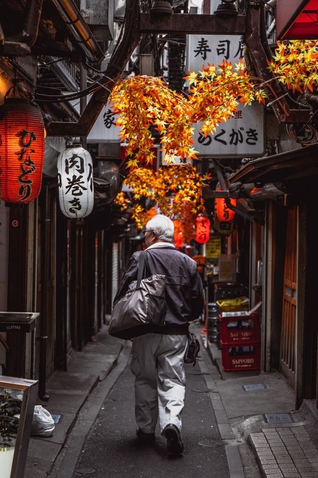 an old man walks down a narrow alley way, decorated with laterns and leaves changing colors, in Tokyo