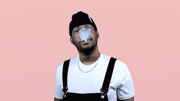 A man in a white shirt, black overalls, and black beanie blows smoke out from his mouth