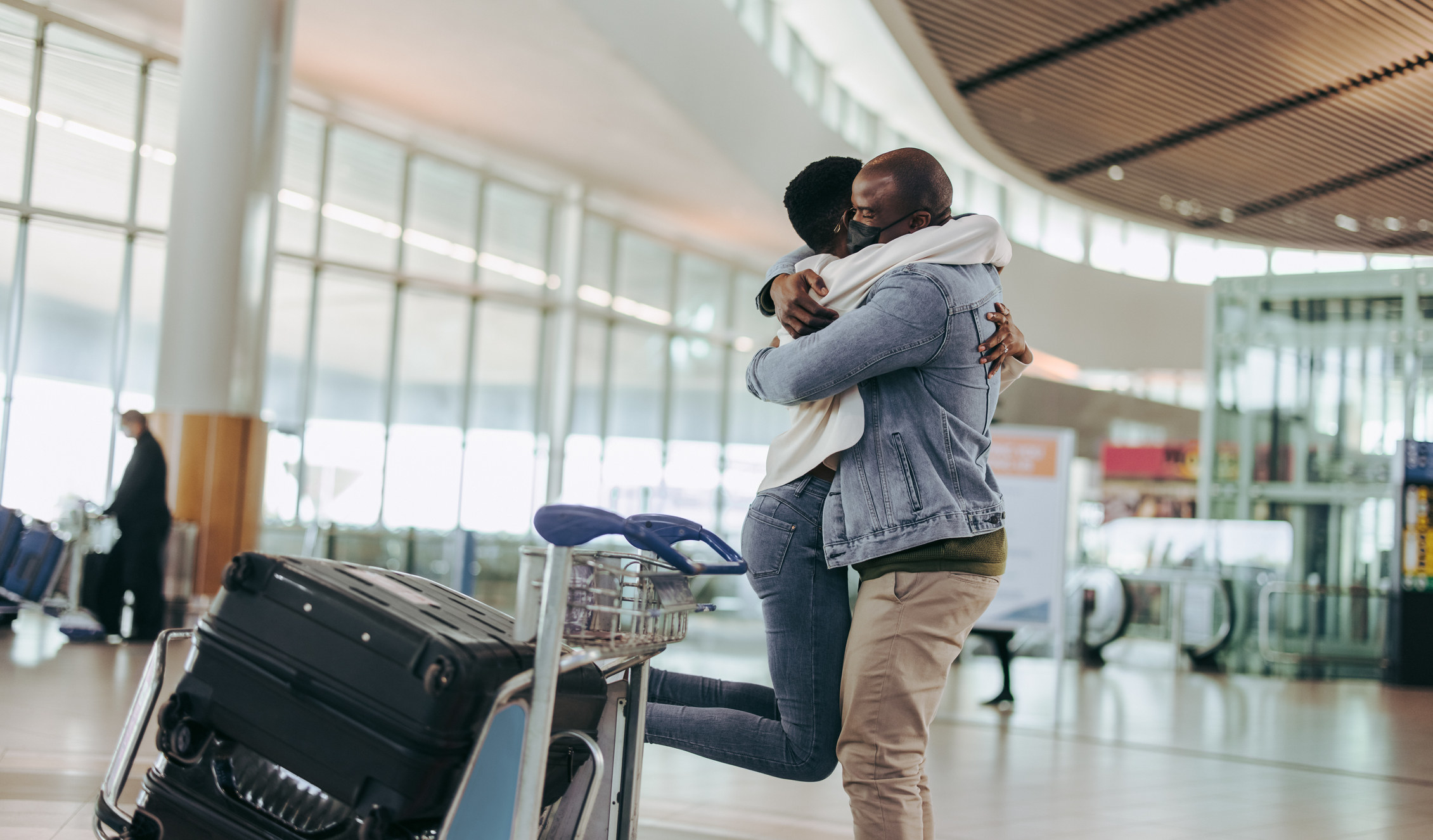 Man with face mask embracing and lifting his girlfriend at airport terminal