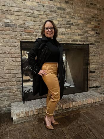 reviewer posing in front of a fireplace where beige leather pants