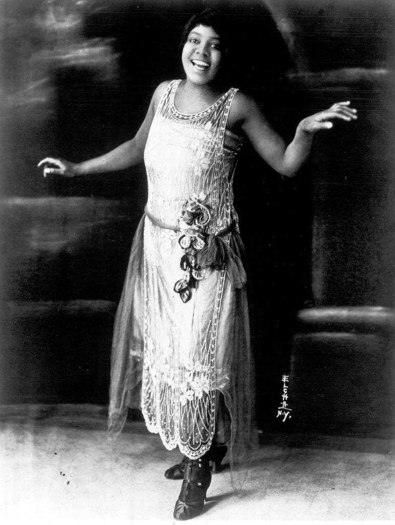 A Black woman smiling and wearing a &#x27;20s-style sleeveless dress
