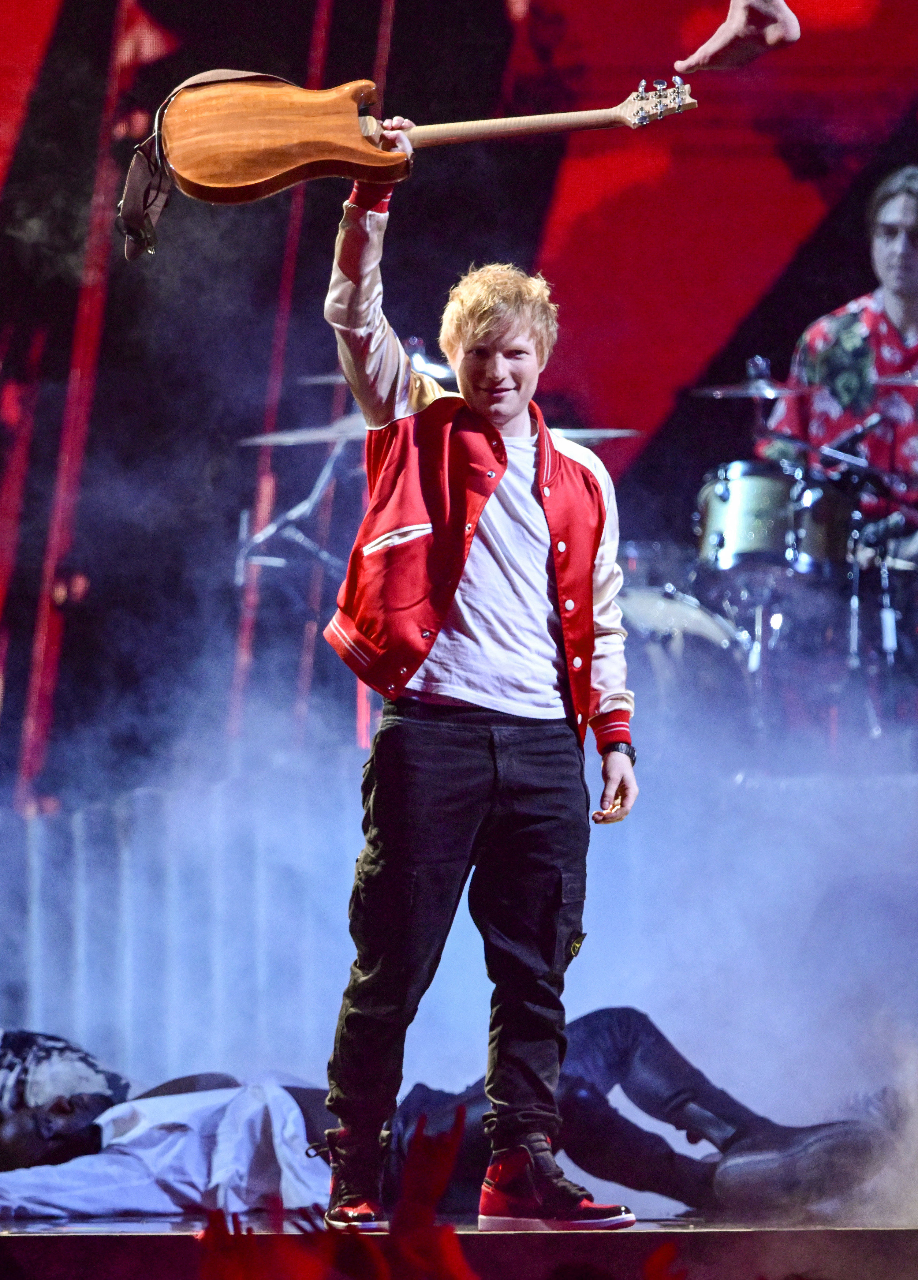 Ed wears a red letterman jacket and holds up his guitar