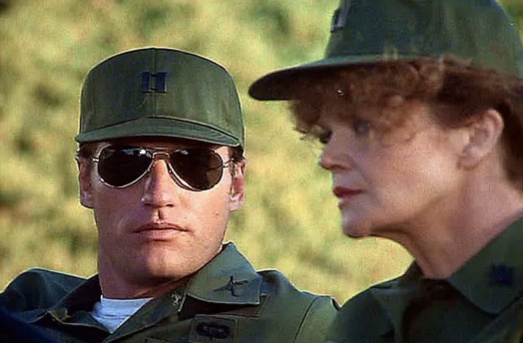 A man in aviator sunglasses looks at a woman. They both wear Army uniforms