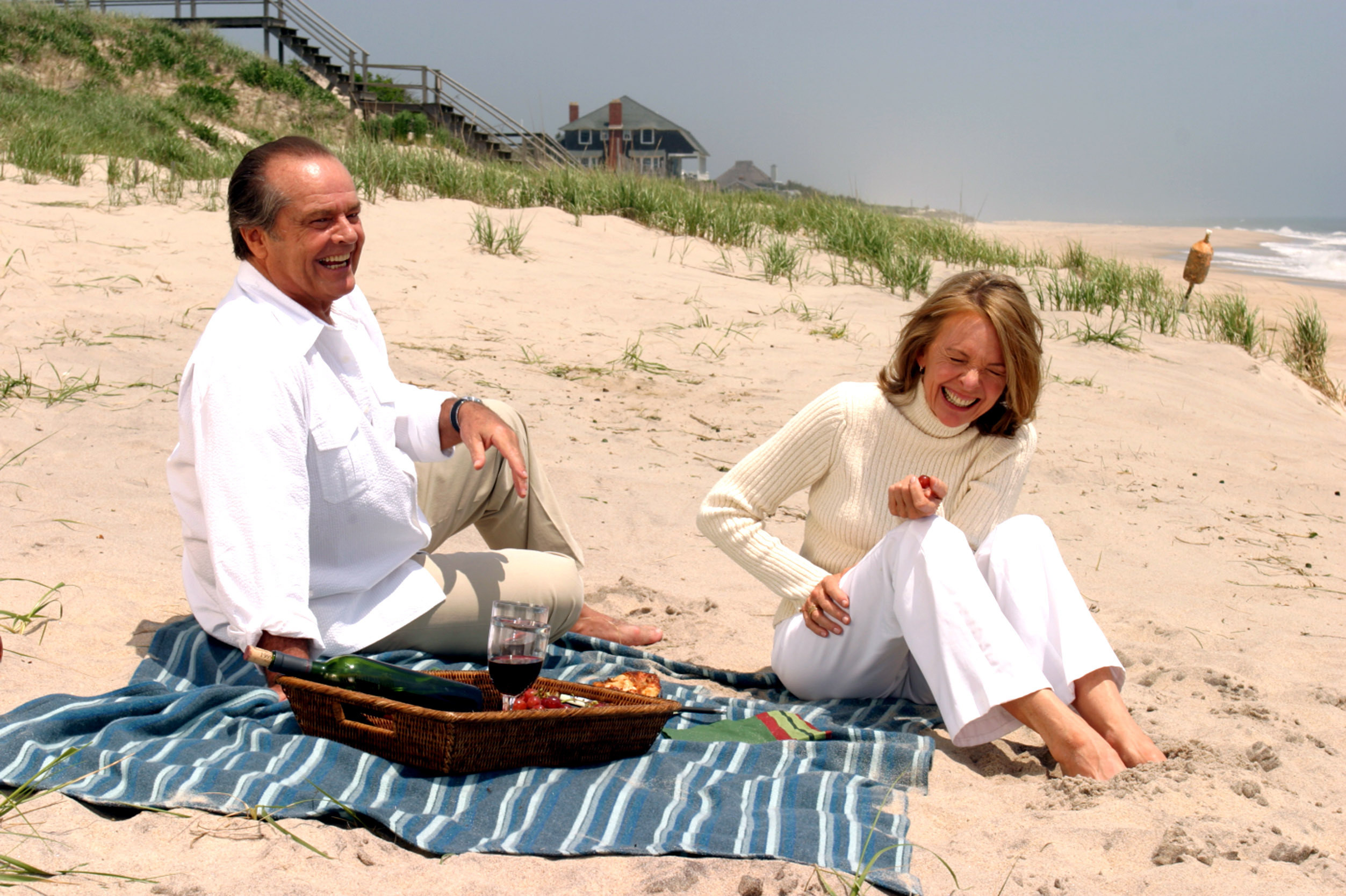 An older man and woman sit on the beach laughing