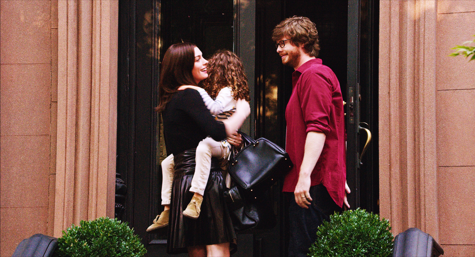 A woman, holding her small daughter, and a man stand in front of a brownstone