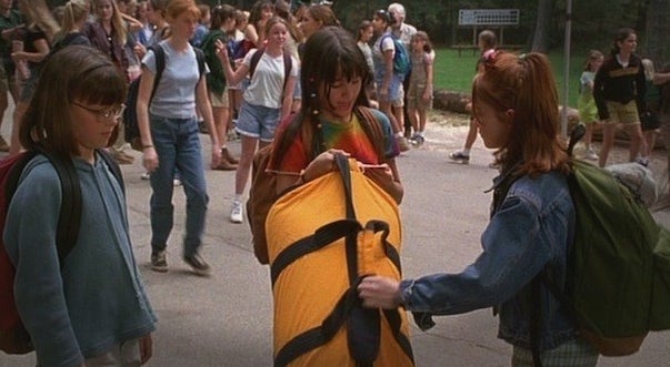 A girl in a tie-dye shirt holds a yellow duffle. A red haired girl reaches for it