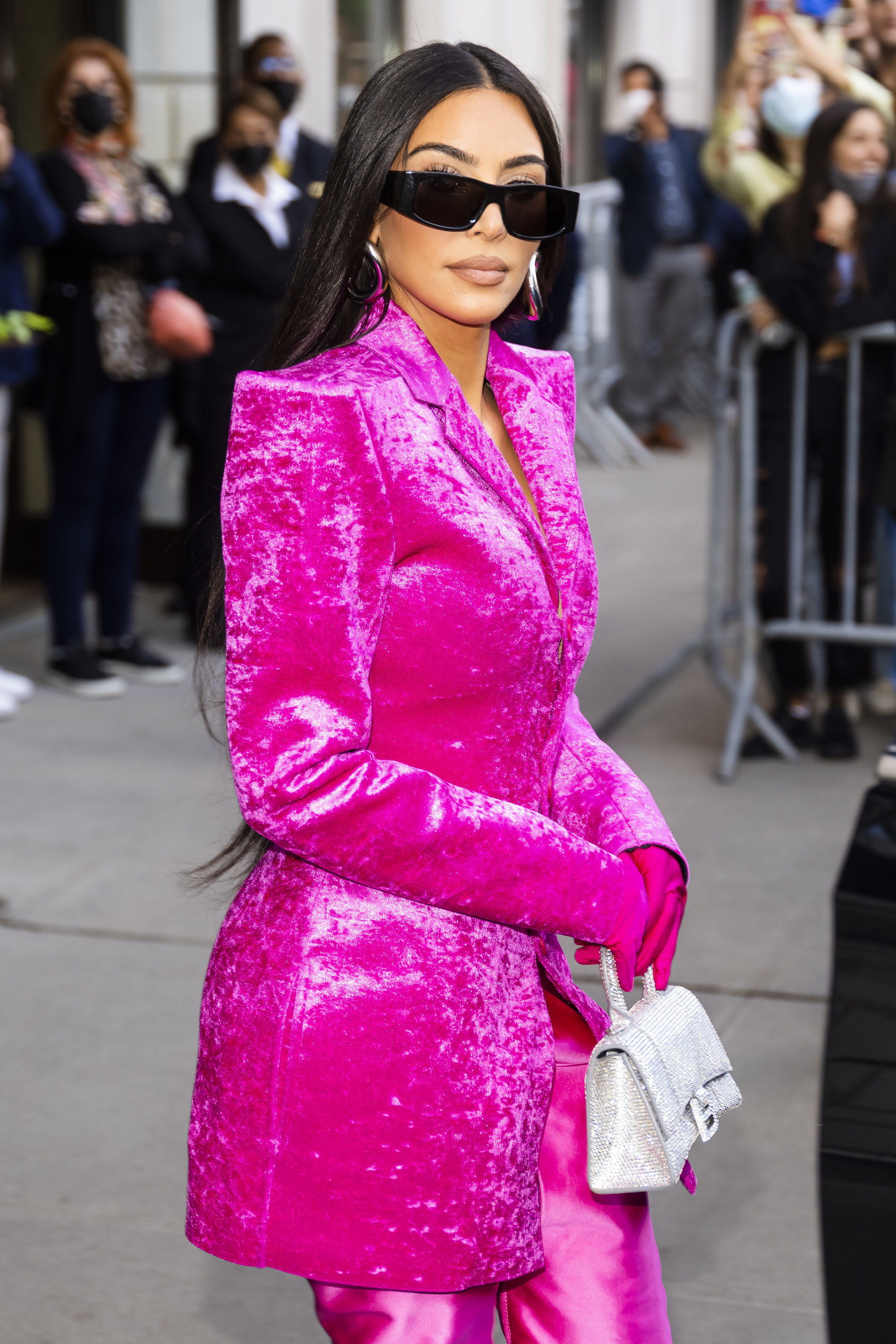 Kim walking out of a building in a long velvet blazer, matching pants, gloves, oversized glasses, and holding a small bejeweled purse