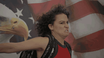 Ilana Glazer from Broad City saluting and looking serious with a US flag behind her
