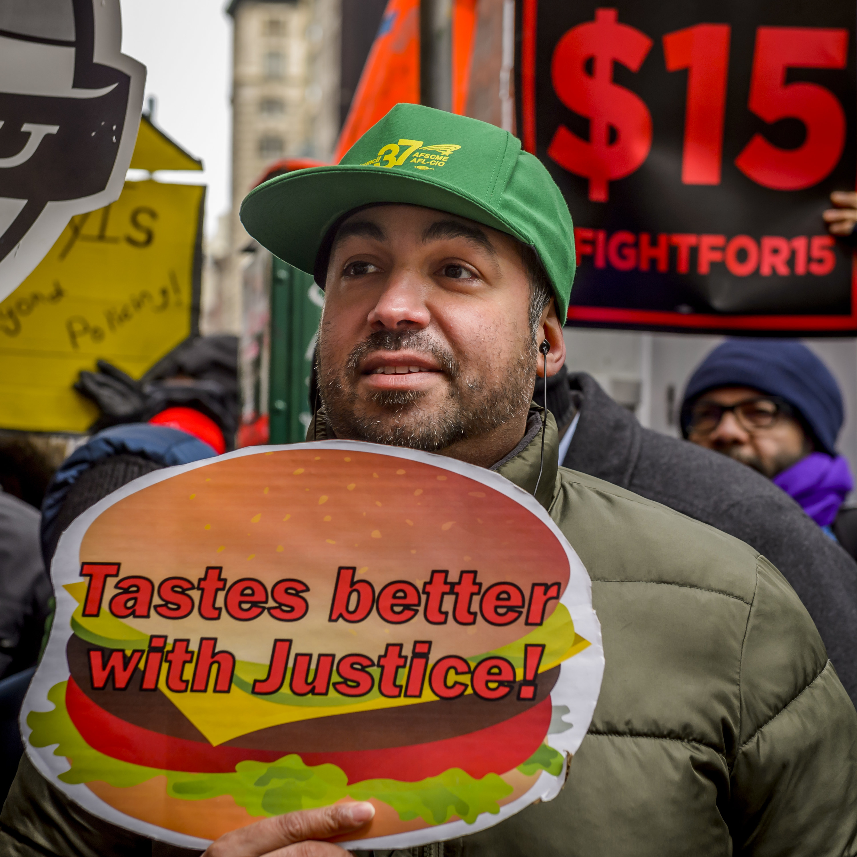 Man at a rally for raising the minimum wage holding a sign shaped like a burger that says tastes better with justice