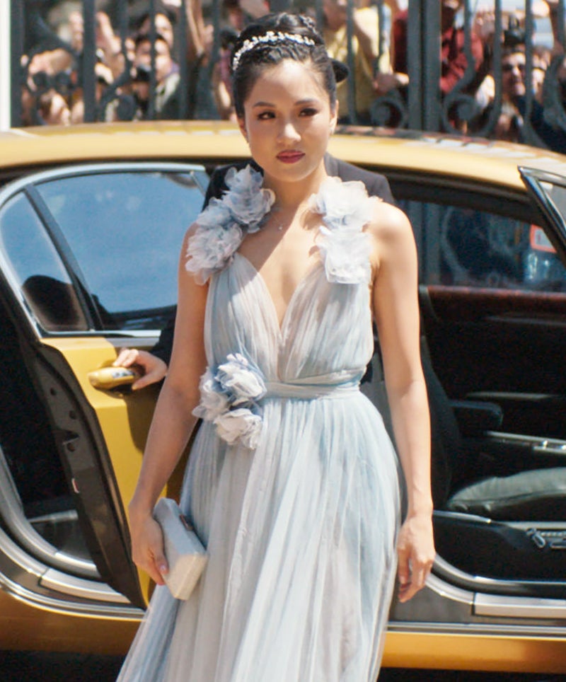 Rachel stands by a car wearing a long, flowing dress with floral accents