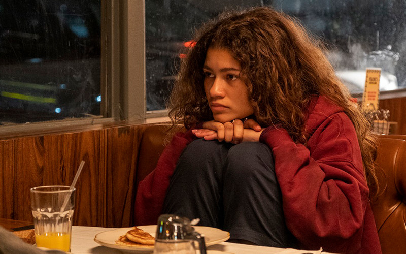 Zendaya playing her character &quot;Rue&quot; in Euphoria is seen in a café with her knees to her chest and her hands on top staring out the window
