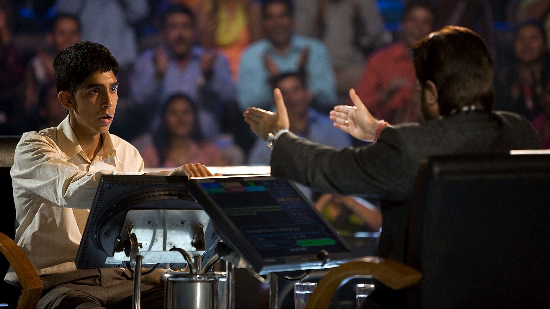 Dev Patel as Jamal Malik sitting in a game show chair as the host Prem Kumar, played by Anil Kapoor, asks him a question