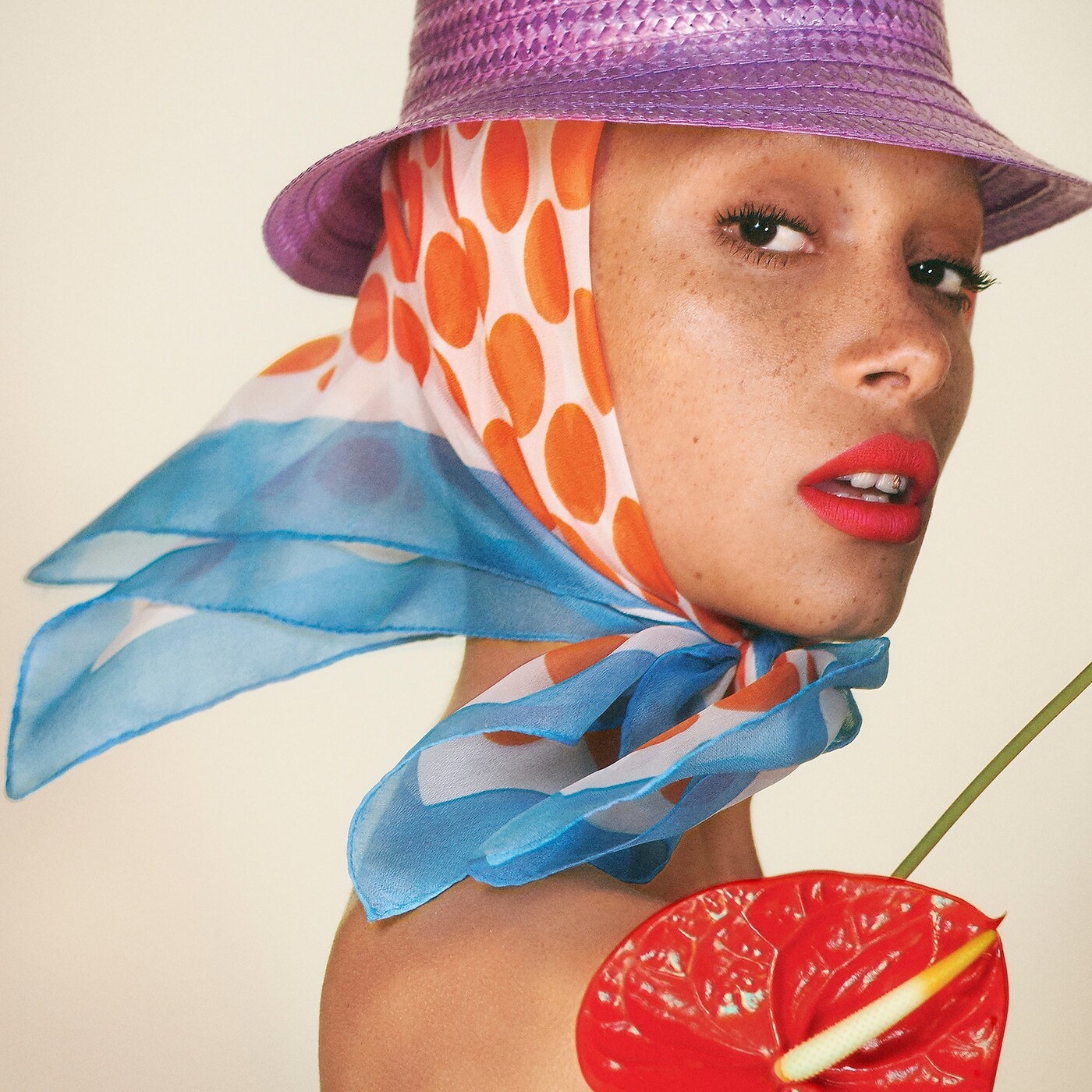Model wearing red lipstick and blue, orange and white scarf with purple hat holding red flower