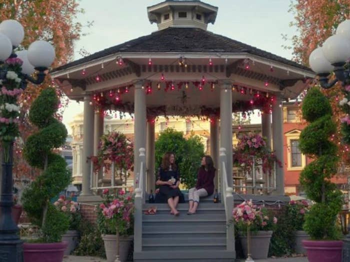 Lorelai and Rory from Gilmore Girls sit on the top step under a gazebo that&#x27;s decorated with lights, town buildings are behind them
