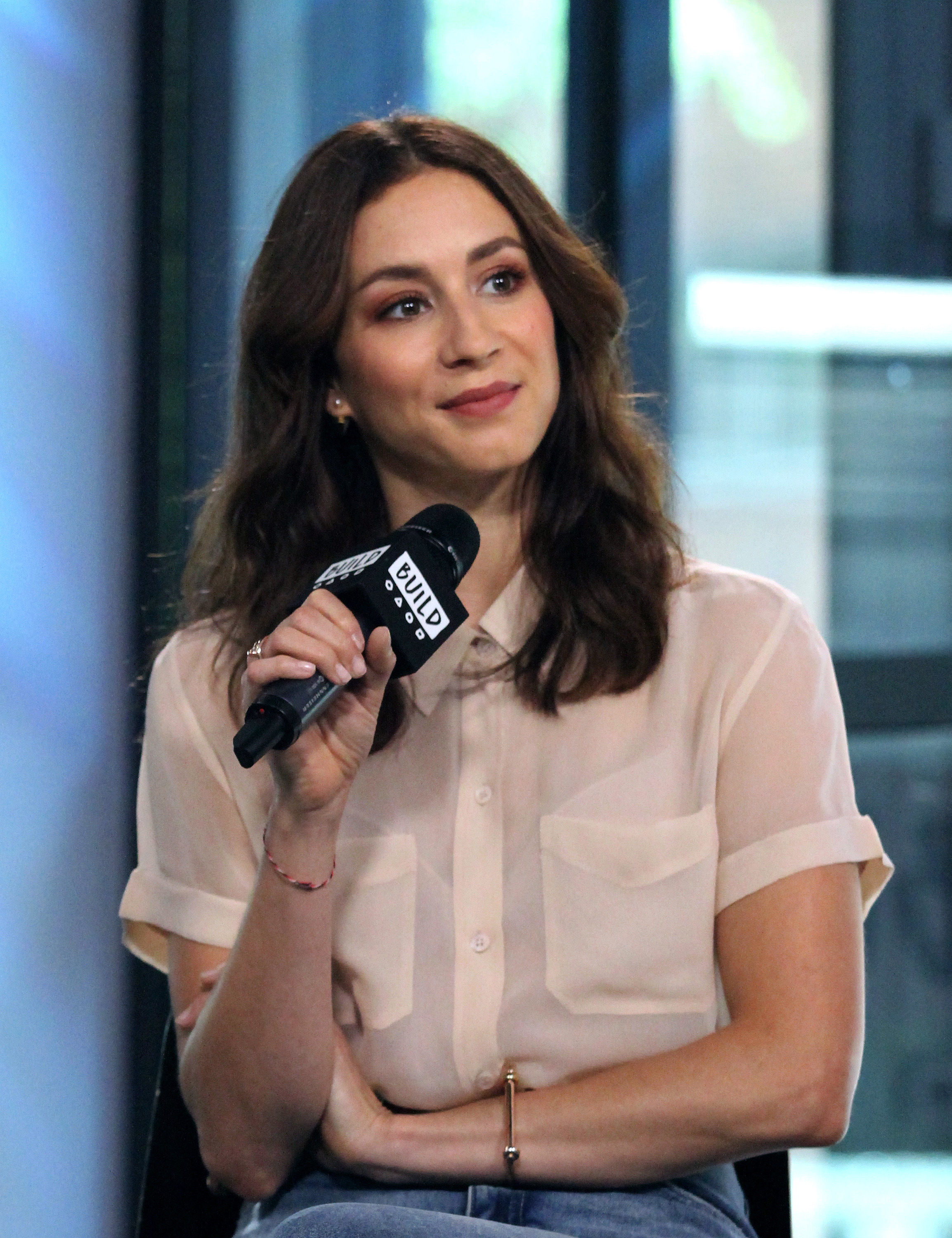 Troian Bellisario sits in a chair holding a microphone, wearing a pale pink shirt