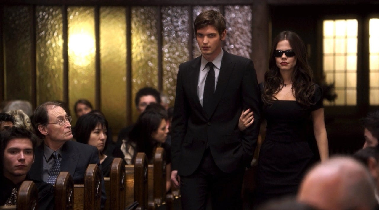 A man in a suit is walking down a church aisle with Jenna, who is dressed in black and wearing dark sunglasses