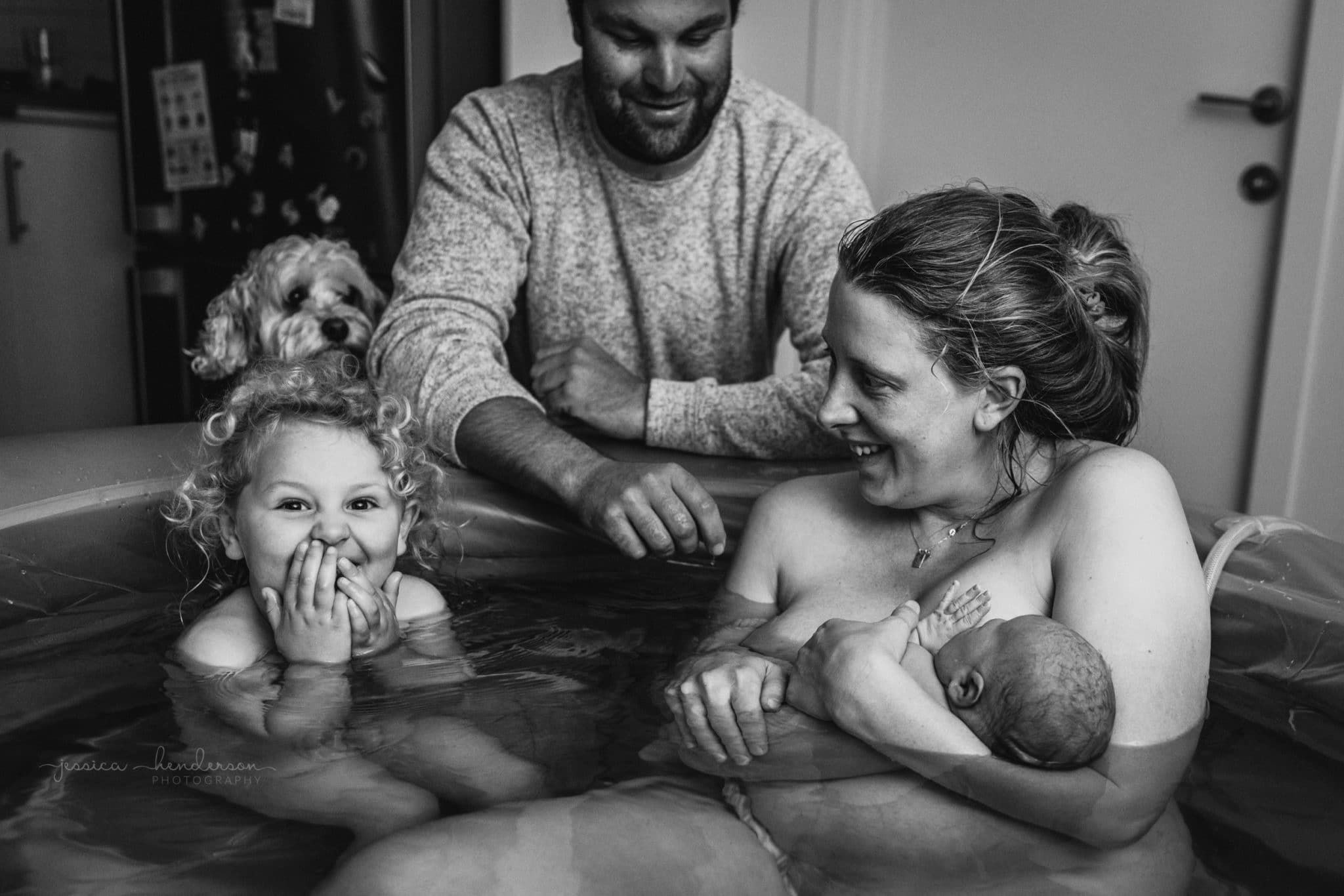 A awestruck, smiling toddler sits in the pool post water birth with her mom and new baby