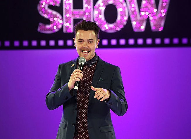 Ray Quinn on stage in a blue suit with a dark red patterned shirt. He has a mic in his hand