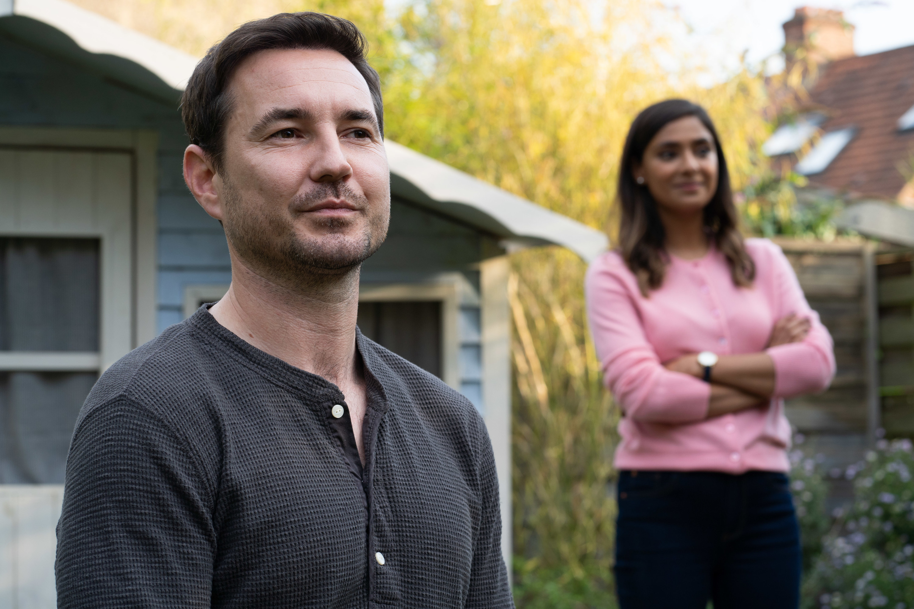Martin Compston as Bram Lawson and Dinita Gohil as Lucy Vaughan standing in the garden, both with wry smiles