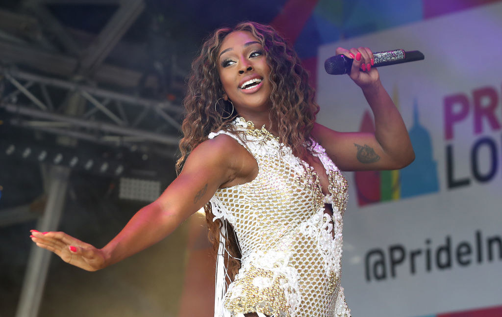 Alexandra Burke perform at London Pride, with a microphone in hand. She is wearing a gold and white bodysuit