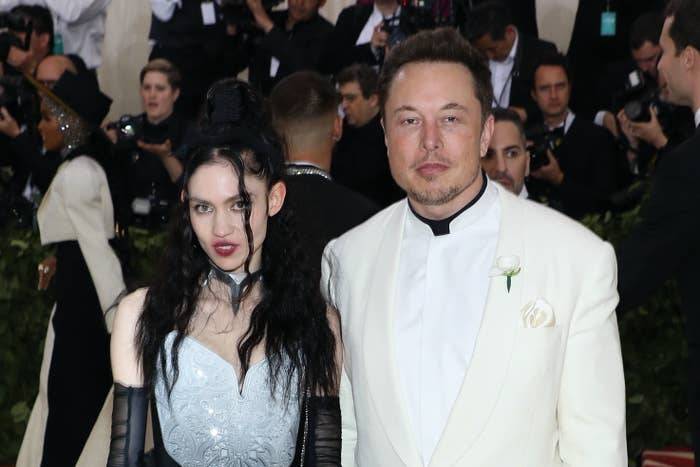 Grimes wears vampire-esque makeup, long transparent gloves and a dress next to Elon, who is wearing a suit with a collar-less shirt