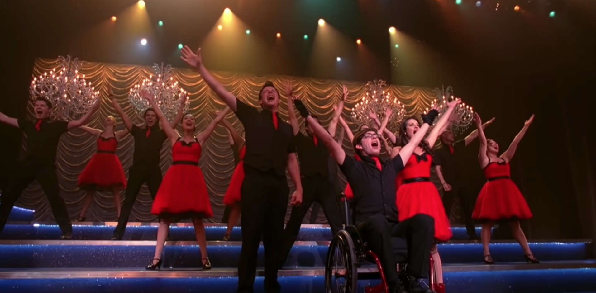 The Glee club performs on stage as the girls wear brightly colored dresses and the guys wear dark button up shirts with brightly colored ties