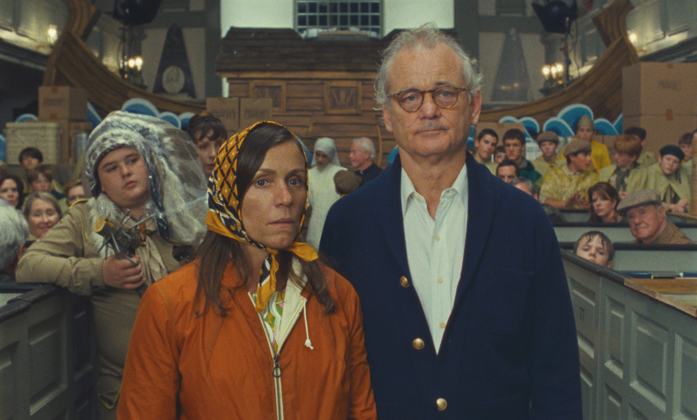 A woman, Frances McDormand, with a scarf tied around her head and a man, Bill Murray, wearing glasses and a cardigan, looking unimpressed