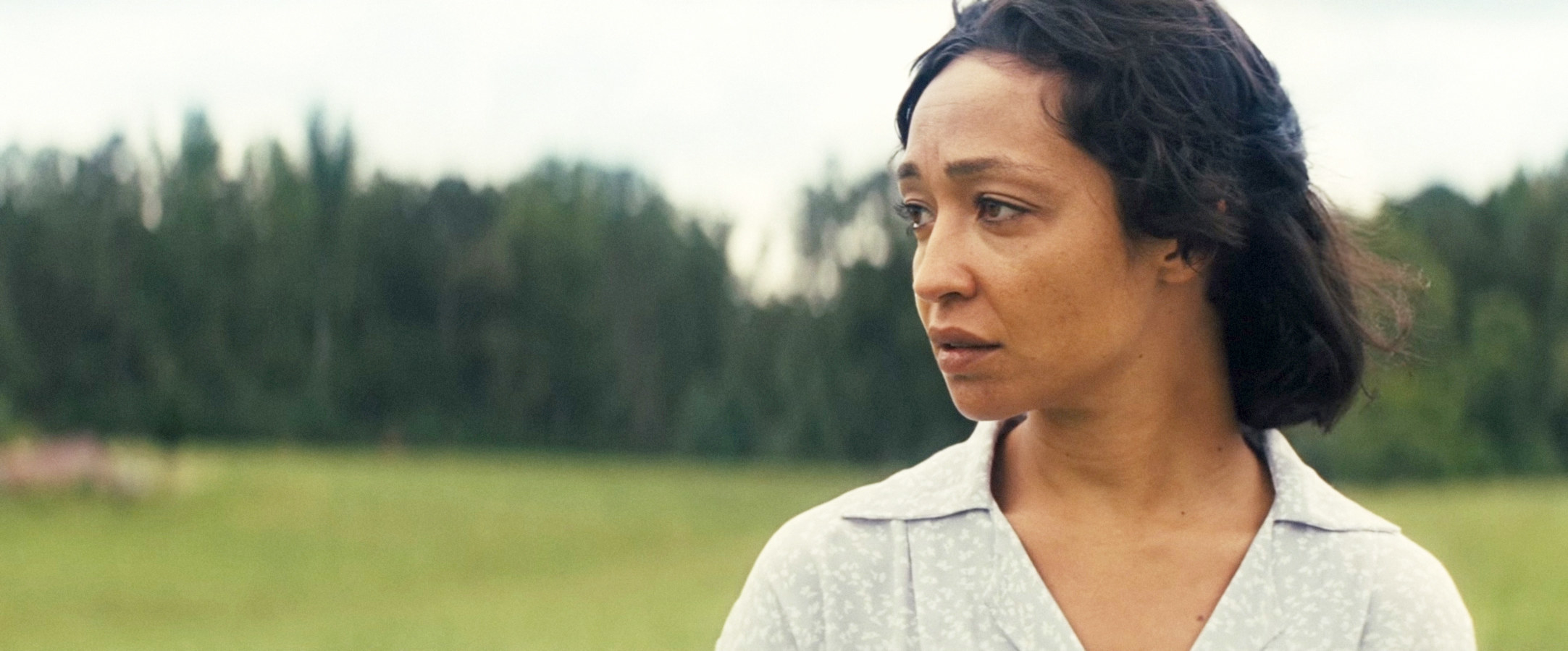 Ruth Negga with her hair tied back, in a house dress, looking out over her shoulder standing in a field