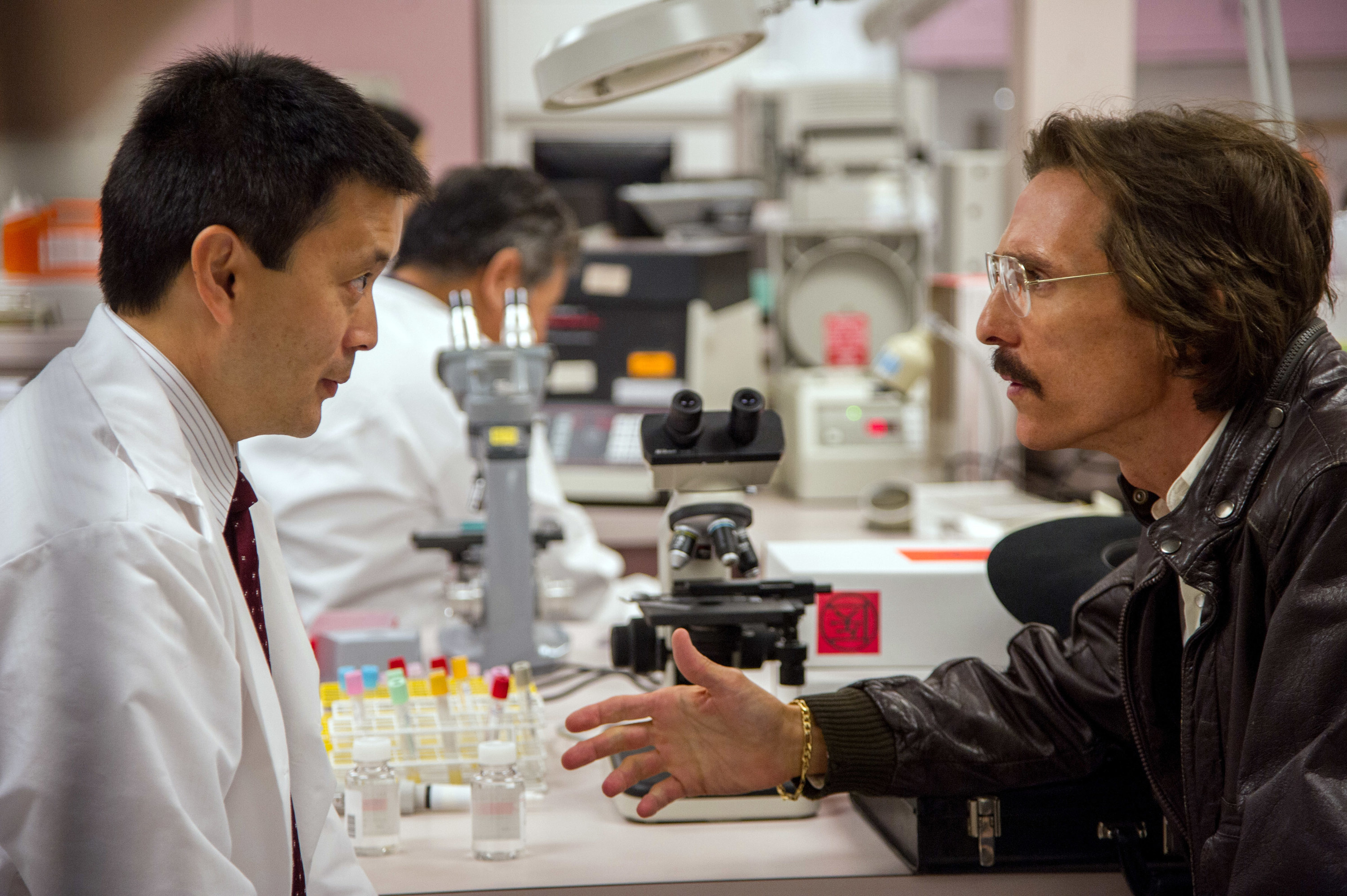 Matthew McConaughey in character wearing a leather jacket, talking to a lab technician
