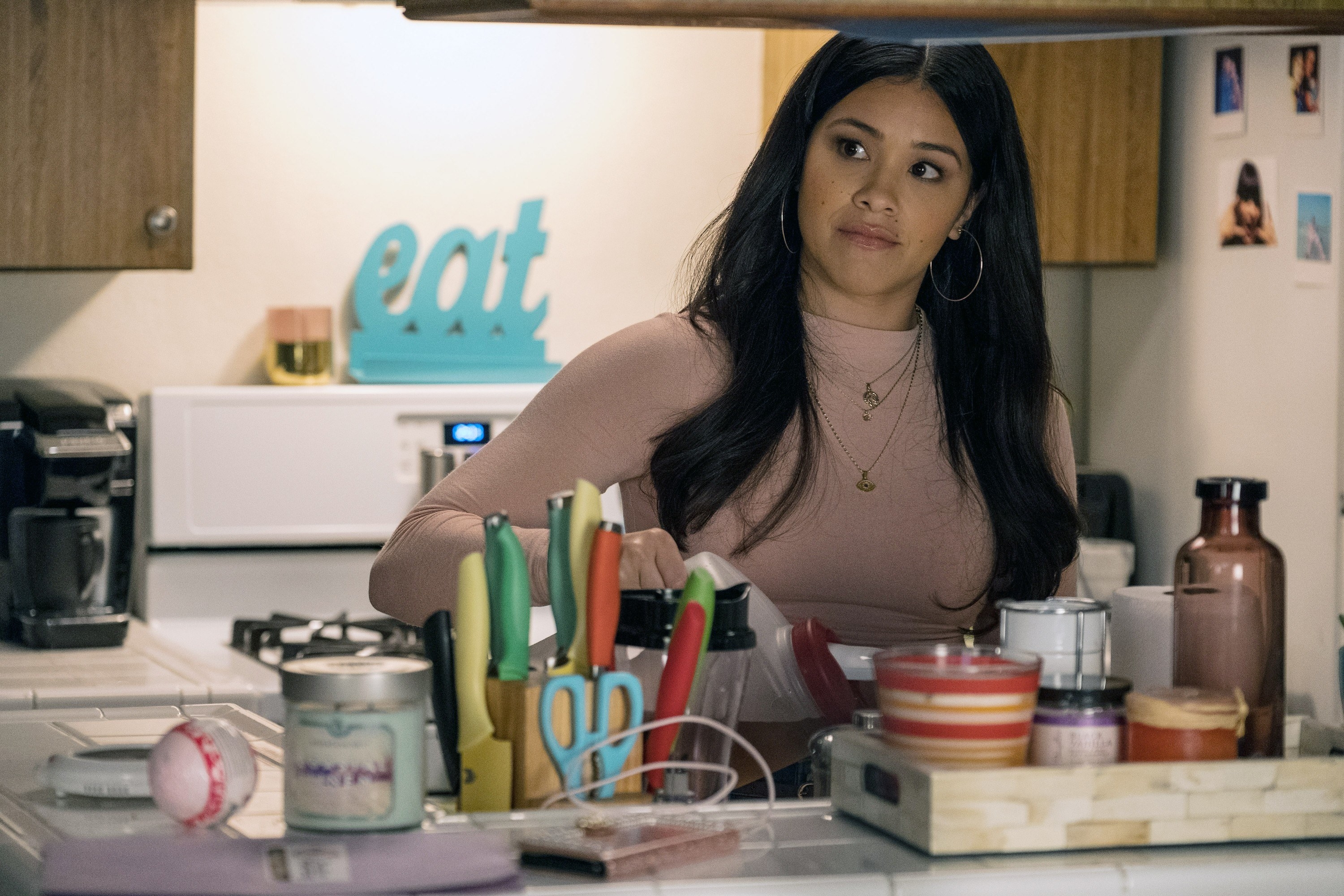 Gina Rodriguez with long hair and a pink top, pouring a drink while looking out through her modest kitchen
