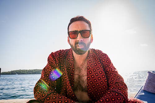 Nicolas Cage in a robe and sunglasses sitting on the beach in the sun.