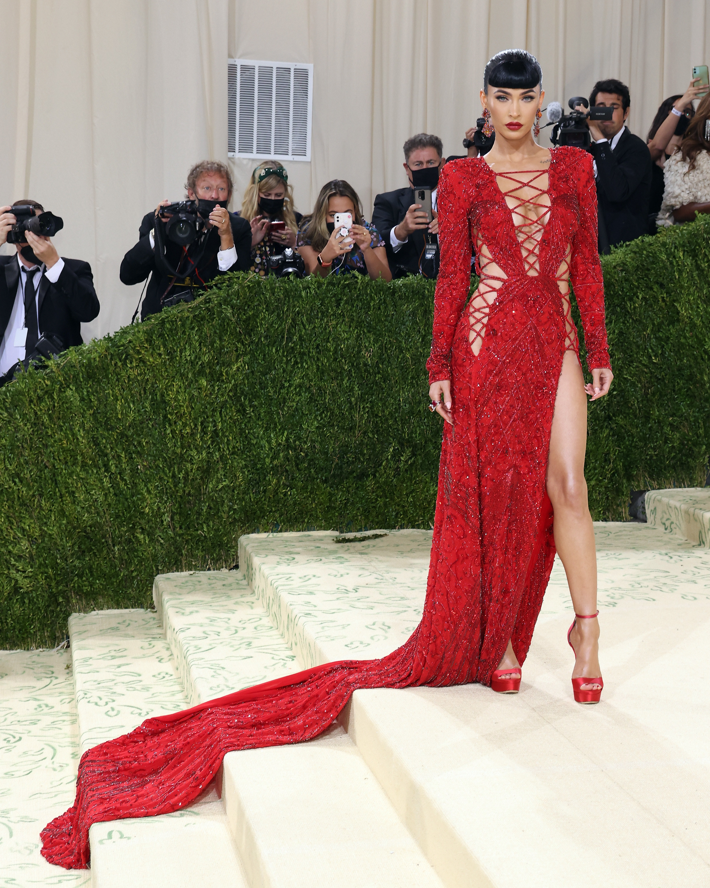 Megan wears a red gown with cutouts and criss-cross details across her body