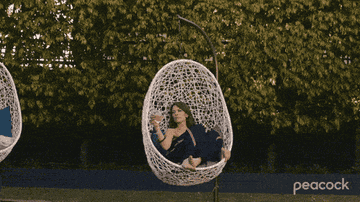 GIF of actor in the show &quot;Five Bedrooms&quot; sitting in a white egg chair drinking wine