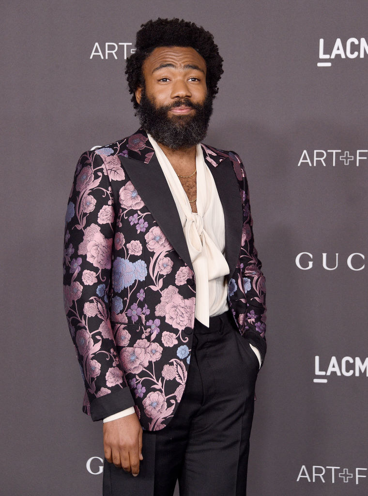 wearing a shiny floral suit jacket, low-cut shirt with a bow, and trousers, Donald Glover smiles on the red carpet