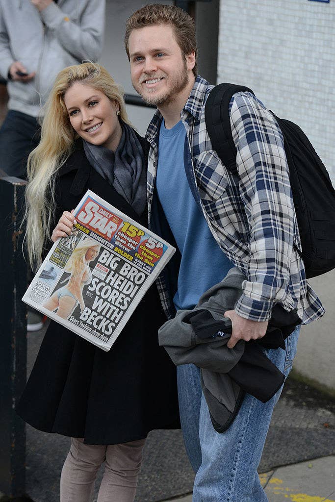 Heidi and Spencer smiling for the paparazzi as Heidi holds up a tabloid with her wearing a bikini on the cover